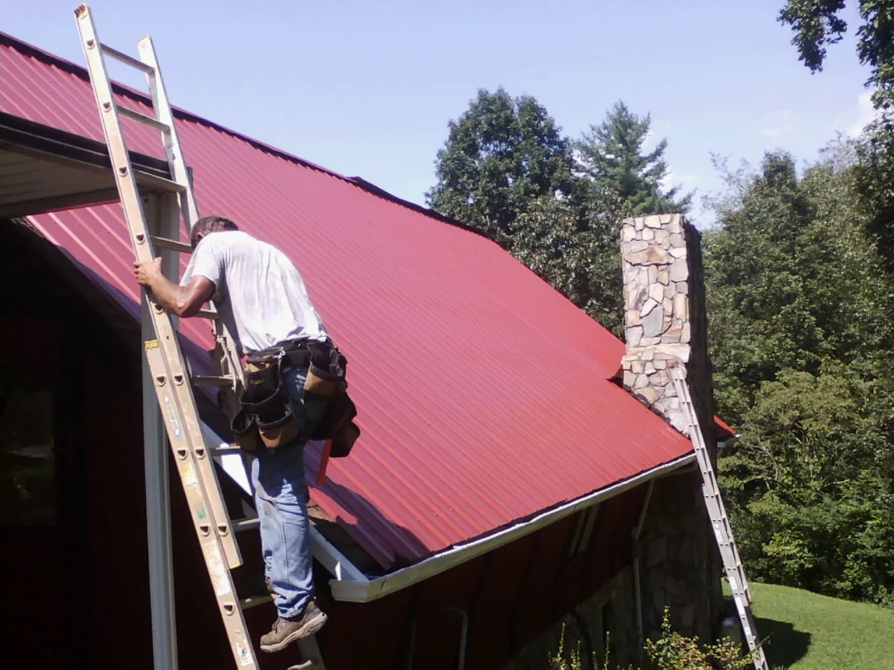 Roofer on a ladder beside a red metal roof and stone chimney — Soffit & Fascia Repair by Roofing Brothers in Cherry Tree